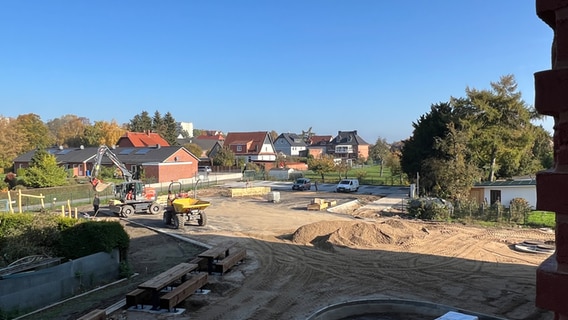 Die Baustelle des Außenbereichs des neuen Grundschulzentrums in Boizenburg. Ein Bagger und ein Radlader stehen auf der brachen, umzäunten Fläche, in deren Mitte sich ein Berg mit aufgeschütetter Erde türmt. Mehrere Holzbänke mit Tischen sind schon installiert und im planierten Boden verankert. Verschiedene Baumaterialien liegen am Rand der Fläche. Im Hintergrund ragen Einfamilienhäuser mit größeren Gärten in den blauen Himmel. © NDR Foto: Lucie Kluth