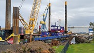 Kran, Bagger und anderes Baugerät baut an einer Wasserspundwand. © NDR Foto: Christoph Woest