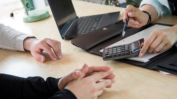 In einer gestellten Szene sitzen zwei Menschen an einem Tisch einer dritten Person gegenüber, die ihnen etwas auf einem Taschenrechner zeigt. Auf dem Tisch steht ein Laptop, vor der dritten Person liegt außerdem eine aufgeklappte Arbeitsmappe. Von allen Beteiligten sind nur die Hände zu sehen. © picture alliance / dpa-tmn Foto: Christin Klose