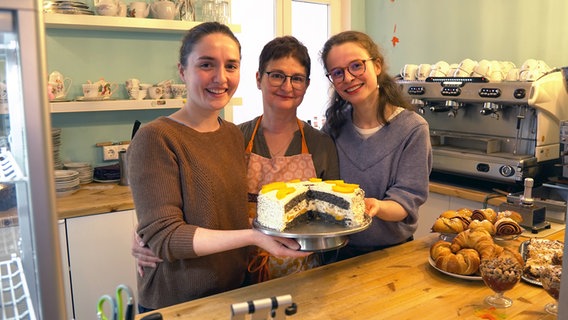 Drei Frauen halten lächelnd eine Torte in die Kamera. Sie stehen dabei hinter einem Tresen in einem Café. © NDR Foto: Christoph Woest