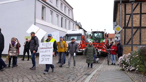 Demonstrierende Anwohner gehen friedlich an einem LKW und einem Traktor vorbei, die eine enge Straße versperren. © NDR Foto: Christoph Woest