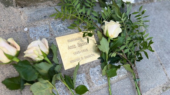 Ein Stein aus Messing in einen Gehweg eingelassen, daneben liegen Rosen. Auf dem Stein steht: Kiebitzberg 1, Anna Becker 1883-1942, ermordet in Auschwitz. © NDR Fotos Foto: Jürn-Jakob Gericke