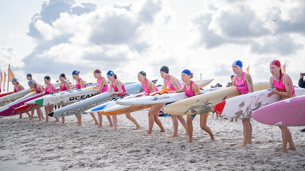 13 Rettungsschwimmerinnen stehen vor sonnig-wolkigem Himmel in einer Reihe am Strand. Sie tragen pinke Badeanzüge mit dem DLRG-Logo und Badekappen. Offensichtlich beginnt gleich ein Rennen - die Frauen befinden sich in lauernder Stellung und haben ihre Surfbretter unter den Arm geklemmt.
