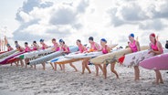 13 Rettungsschwimmerinnen stehen vor sonnig-wolkigem Himmel in einer Reihe am Strand. Sie tragen pinke Badeanzüge mit dem DLRG-Logo und Badekappen. Offensichtlich beginnt gleich ein Rennen - die Frauen befinden sich in lauernder Stellung und haben ihre Surfbretter unter den Arm geklemmt. © DLRG Foto: DLRG