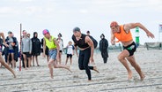 Drei Männer in Badeklamotten liefern sich ein Rennen am Strand. Die Anstrengung steht ihnen in die Gesichter geschrieben - unter vollem Einsatz preschen die Athleten über den Sand. © DLRG Foto: DLRG