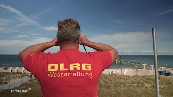 Ein Mann mit einem roten DLRG-Shirt schaut mit einem Fernglas aufs Wasser. © NDR 