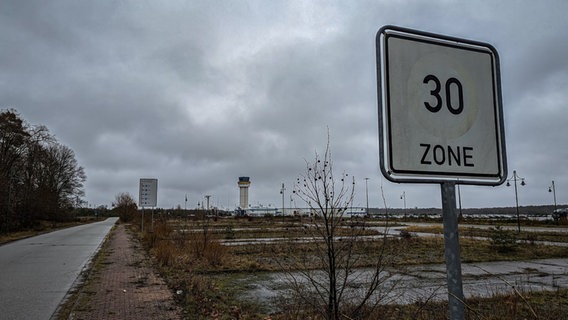 Ein ausgeblichenes 30er-Zonen-Schild, dessen rote Umrandung kaum noch erkennbar ist, zeigt die Geschwindigkeitsbegrenzung auf der Zufahrt zum Flughafenparkplatz an. Im Hintergrund sind das Gebäude, der Tower und der wolkenverhangene Himmel zu sehen. © NDR Foto: Christoph Loose