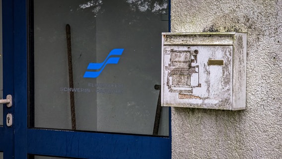 Ein verwitterter Briefkasten an einem Nebengebäude wurde offenbar mehrfach mit unterschiedlichen Namenszügen überklebt. Daneben sind durch die Glastür einige Putzutensilien erkennbar, die im Vorraum herumstehen. © NDR Foto: Christoph Loose