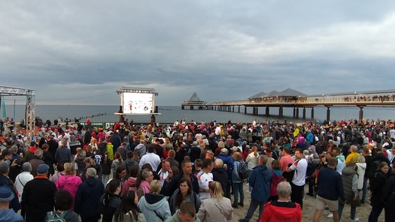 Eine Menschenmenge sieht sich am Strand von Heringsdorf auf Usedom das Auftaktspiel der EM 2024 auf einer großen LED-Videowand an. Im Hintergrund ist das Meer und die beleuchtete Seebrücke zu sehen. © NDR Foto: Steffen Baxalary