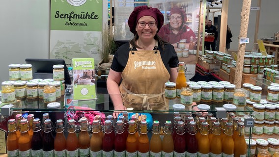 Eine Frau mit Brille, einer Schürze und einer Kochmütze steht hinter einer gläsernen Vitrine an einem Stand der Grünen Woche in Berlin. In und auf der Vitrine stehen zahlreiche Flaschen und Gläser mit Senfkreationen aller Art, allesamt mit dem grünen Label der Senfmühle Schlemmin. Im Hintergrund stapeln sich weitere Gläser. © NDR Foto: Franziska Drewes