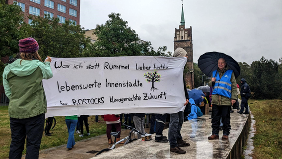 Auf der Grünfläche vor dem Kröpeliner Tor Center stehen Mitglieder des BUND mit einem Transparent. Darauf steht: "Was ich statt Rummel lieber hätte: lebenswerte Innenstädte für Rostocks klimagerechte Zukunft". © NDR Foto: Carolin Beyer