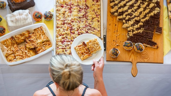 Kuchenbasar von oben: Eine Frau hält einen Teller mit Kuchen über einen mit verschiedenem Gebäck vollgestellten Tisch. © IMAGO / Thomas Müller Foto: IMAGO / Thomas Müller