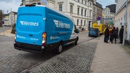 Ein Transporter des Lieferdienstes Hermes mit der bekannten weiß-blauen Farbe steht am Straßenrand in der Schweriner Arsenalstraße. Daneben eine Gruppe von Menschen, im Hintergrund steht ein weiterer Transporter des Lieferdienstes DHL. © NDR Foto: Christoph Loose