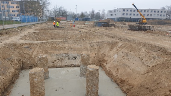 Eine große, komplett eingezäunte Baustelle zieht sich über den ehemaligen großen Parkplatz am Lambrechtsgrund in Schwerin. Das gesamte Erdreich wurde ausgehoben und tiefe Löcher gegraben. In eines der Löcher wurden bereits vier massive, rundliche Betonpfeiler gerammt, dann wurde es zum Teil mit Beton ausgefüllt - die Pfeiler ragen aus einer ebenen, grauen Masse hervor. Im Hintergrund sind Bauarbeiter, Container, Erdhügel, Metallstreben, Messgeräte und verschiedene Baumaschinen zu sehen. Hinter der Umzäunung sind ein Fußballplatz und ein modernes Kastengebäude zu sehen. © NDR Foto: Wiebke Heiden