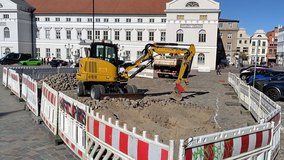 Ein gelber Bagger steht auf dem Wismarer Markt. Um ihn herum ist ein Bereich mit einem Bauzaun eingezäunt. Ein Loch im Pflastersteinboden ist ausgehoben. © NDR Foto: Christoph Woest