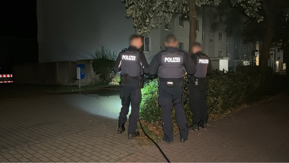 Drei Polizisten stehen in Uniform im Dunkeln, von der Kamera abgewandt an einer kleinen Grüninsel neben einem Gehweg und betrachten den Tatort eines Messerangriffs. © Felix Baenz Foto: Felix Baenz