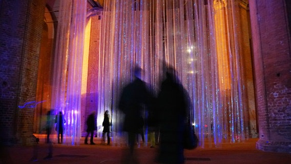 In einer Kirche hängen lange Elemente herunter, die im Dunkeln leuchten. Davor steht ein Paar, welches sich die Ausstellung anschaut. © NDR Foto: Christoph Woest