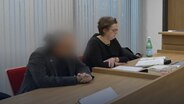 Ein Mann sitzt auf einer Anklagebank. Sein Gesicht ist verpixelt. Daneben sitzt seine Verteidigerin. © NDR 