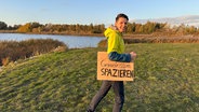 Ein Mann steht mit einem Pappschild mit der Aufschrift "Gemeinsam Spazieren" an einem See. © NDR Foto: Jan Tauer