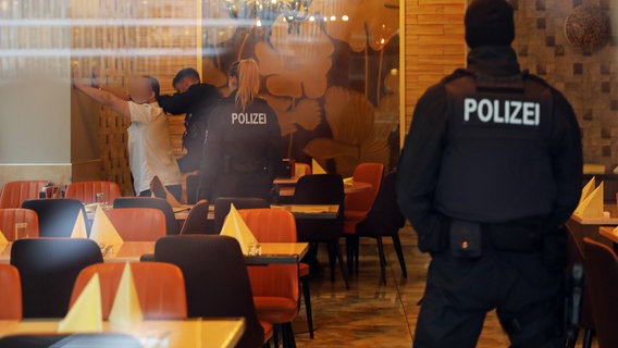 Durch eine Scheibe fotografiert stehen Polizisten von hinten zu sehen in einem Restaurant. An eine Wand gelehnt wird ein Mann durchsucht. © Stefan Tretropp Foto: Stefan Tretropp