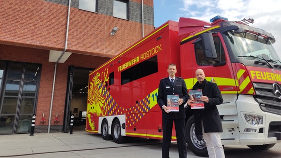 Michael Allwardt und Chris von Wrycz Rekowski halten den Jahresbericht der Feuerwehr in der Hand. Hinter ihnen der neue Einsatzleitwagen. © NDR Foto: Laureen Zielke