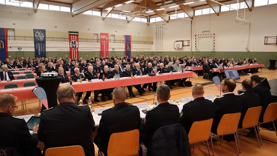 In einer Turnhalle in Neubukow sitzen an langen Tischreihen Feuerwehrleute in Uniform © NDR 