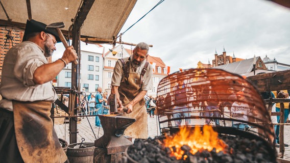 Zwei Schausteller stehen mit Hammer neben einer Feuerschale und schmieden ein glühendes Eisen. © Hansestadt Stralsund Foto: Michelle Dynio