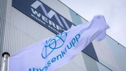 Die Fahne des Unternehmens ThyssenKrupp weht vor der Schiffbauhalle auf dem Gelände der Werft in Wismar, an der noch der Schriftzug MV Werften zu sehen ist. © dpa Bildfunk Foto: Jens Büttner