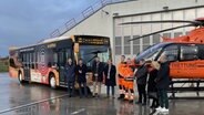 Rebus-Chef Thomas Nienkerk, Landrat des Landkreises Rostock Sebastian Constien (SPD), und weitere Personen stehen in einer Reihe vor einem Bus und einem Rettungshubschrauber. Sie präsentieren einen der ersten Busse des Unternehmens, die mit einem Defibrillator ausgestattet sind. © NDR Foto: Marian Thürmer