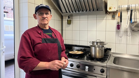 Der Rentner und Koch Peter Brietzke an seinem Arbeitsplatz. Er trägt eine Brille, eine blaue Kappe der Marke Puma und eine rote Schürze. Hinter ihm brutzeln Schnitzel in einer größeren Pfanne unter einem imposanten Abzug. Auf dem Herd stehen außerdem zwei Töpfe. Daneben ist eine Spüle zu sehen, über der verschiedene Küchenutensilien hängen. Der Raum mit weiß gefliesten Wänden ist hell ausgeleuchtet. © NDR Foto: Lucie Kluth