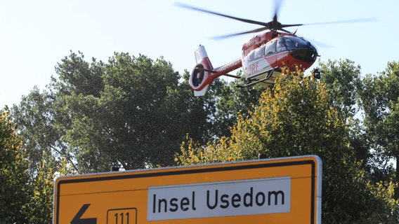 Ein Rettungshubschrauber fliegt in bodennähe an einer Unfallstelle. Im Vordergrund ist ein weißt ein Straßenschild die Richtung zu Insel Usdeom © NDR Foto: Tilo Wallrodt