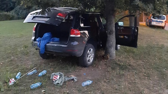 Ein verunfallter VW Touareg steht neben einem Baum auf einer Grasfläche. Aus dem offenen Kofferraum hängt eine blaue Jacke, davor liegen mehrere Plastikflaschen und auch weitere Kleidungsstücke. Im Hintergrund ist ein Polizeiwagen zu sehen. © Bundespolizeiinspektion Pasewalk Foto: Bundespolizeiinspektion Pasewalk