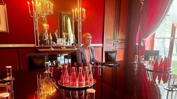 Schwesig in der Residenz des deutschen Botschafters in Paris. Sie sitzt an einem großen Tisch in einem Zimmer mit dunkelroten Wänden. Durch ein bodentiefes Fenster dringt Tageslicht an schweren roten Vorhängen vorbei in den Raum. Auf dem Tisch stehen Wasserflaschen und Gläser, hinter Schwesig hängen Kerzenständer und ein Spiegel, in dem man ihre Begleiter sieht, die noch nicht sitzen. Neben sich auf dem Tisch hat Schwesig eine Mappe liegen. © NDR MV Foto: Anna-Lou Beckmann