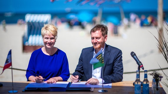Manuela Schwesig (SPD) und Michael Kretschmer (CDU) unterzeichnen Kooperationen, im Hintergrund der Strand von Warnemünde. © dpa-Bildfunk Foto: Jens Büttner/dpa