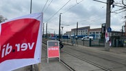 Ein Aufsteller und eine große Fahne der Gewerkschaft ver.di stehen unmittelbar vor dem Betriebshof der RSAG in Rostock. Dort stehen mehrere Straßenbahnen. © NDR Foto: Denis Mollenhauer