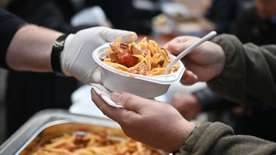 Eine mit Einweghandschuhen behandschuhte Hand reicht eine Plastikschüssel mit Essen, eine andere Person greift mit beiden Händen danach. © picture alliance/dpa Foto: Uli Deck