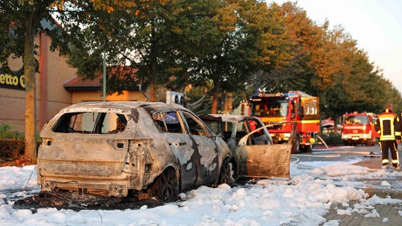 Zwei nahezu komplett ausgebrannte Autos stehen ineinander verkeilt auf der Straße, um sie herum eine Lache von Löschschaum. Im Hintergrund sind Einsatzfahrzeuge der Feuerwehr zu sehen. © NDR Foto: Stefan Tretropp