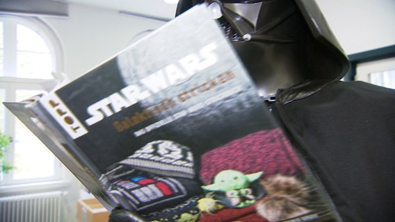 Ein Darth-Vader-Darsteller hält ein Buch über galaktisches Stricken in der Hand. © NDR Foto: NDR
