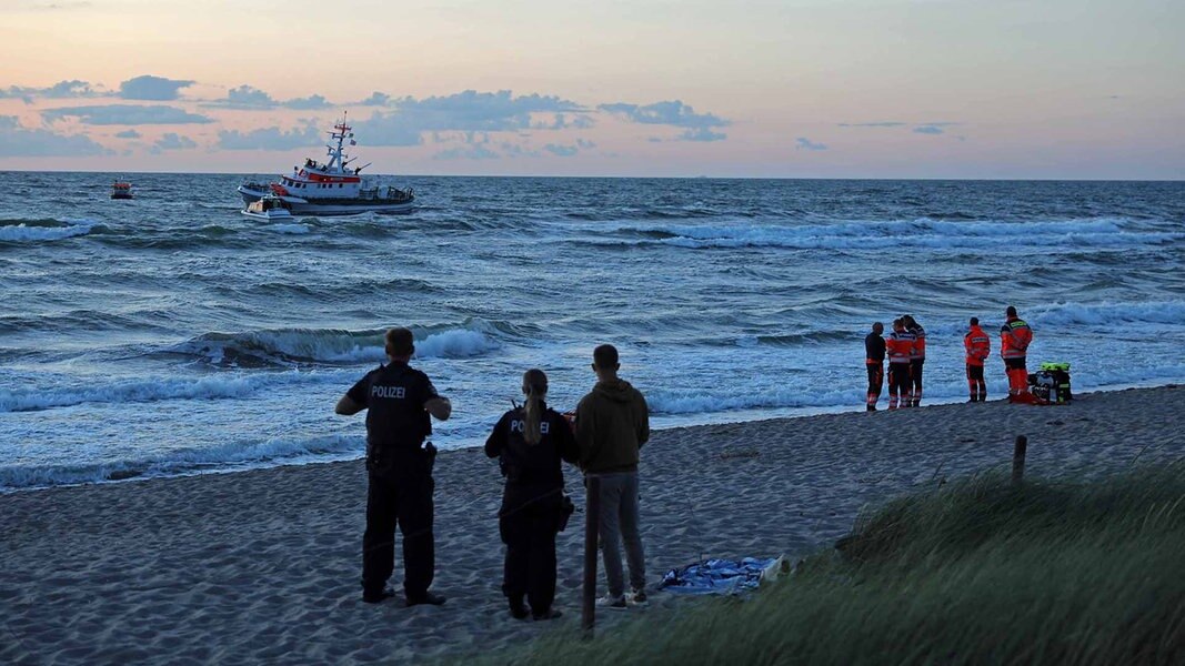 Polizisten und Rettungskräfte stehen in der Abenddämmerung am Strand und suchen die Ostsee nach einem Vermissten ab. Im Wasser in Strandnähe fährt ein Rettungsboot von rechts nach links, ebenfalls auf der Suche. 