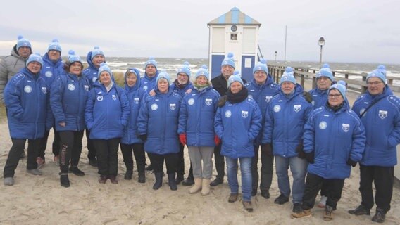 Gruppenfoto des Eisbade Clubs Seehunde Lubmin vor der Seebrücke von Lubmin am Strand. Alle Mitglieder - etwa 20 - tragen die gleichen blauen Jacken mit dem Vereinslogo © Eisbade Club "Seehunde Lubmin" / Harald Köhler 