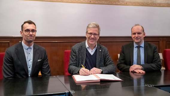 Drei Personen sitzen hinter einem Schreibtisch im Rathaus Greifswald und schauen in die Kamera. © Universitäts- und Hansestadt Greifswald Foto: André Gschweng