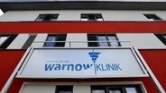 Blick von unten bei blauem Himmel auf die Front der Warnow-Klinik von außen. Ein quadratischer, modern wirkender Bau in rot und weiß, im Vordergrund ein Schild mit dem Logo der Klinik. © dpa Bildfunk Foto: Bernd Wüstneck