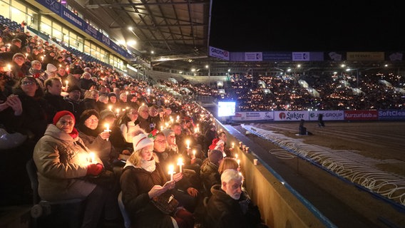 Zahlreiche Menschen halten Kerzen in den Händen und sitzen dick eingepackt im Rostocker Ostseestadion, um gemeinsam Weihnachtslieder zu singen. © picture alliance/dpa Foto: Danny Gohlke