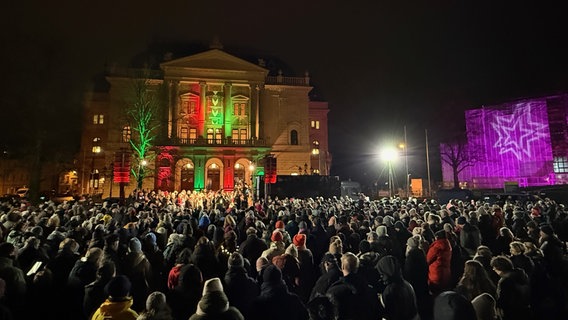 Rund 1.500 Menschen singen im Dezember Weihnachtslieder auf dem Alten Garten in Schwerin. Es ist dunkel und die warm angezogene Menge wendet sich der bunt beleuchteten Fassade des © NDR Foto: Christoph Kümmritz