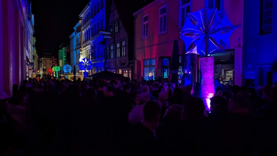 Menschen stehen im Dunkeln in einer Gasse, im Hintergrund und an den Hauswänden bunte Lichter. © Christoph Loose 