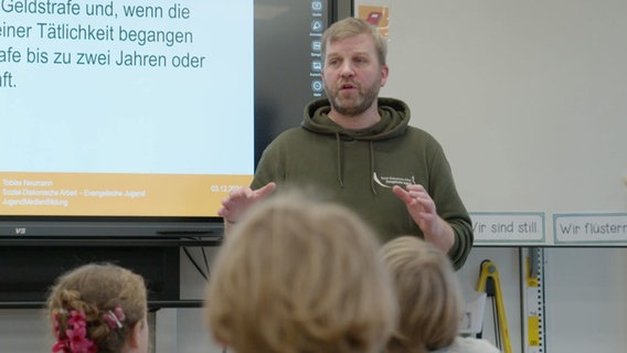 Ein Mann steht in einem Klassenraum vor einer digitalen Tafel. Im Vordergrund sitzen Schüler. © Screenshot 