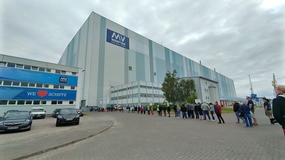 Die Wismarer Werft von außen, davor stehen Menschen in einer Schlange. Foto: Christoph Woest