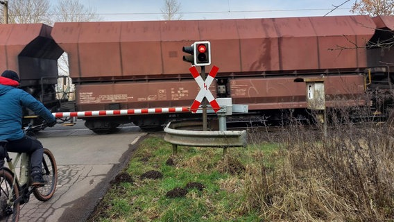 Ein Radfahrer wartet an einem geschlossenen Bahnübergang, während ein Güterzug durchfährt. © NDR Foto: Julia Scheper