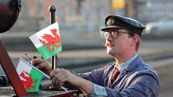 Caleb Lovegrove von der Ffestiniog and Welsh Highland Railways (FR/WHR) aus Wales befestigt an einer Dampflok der Harzer Schmalspurbahnen GmbH die walisische Flagge. © dpa-Bildfunk Foto: Matthias Bein