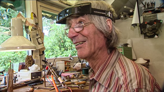 Der Goldschmied Hadfried Rinke arbeitet in seiner Werkstatt an einem Ring und lächelt. © NDR 